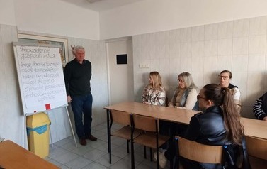 Dwoje dzieci siedzi w kaskach w samochodzie strażackim w kaskach i macha przez przednią szybę.